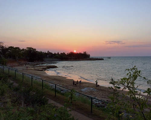 Beautiful sunset over a tranquil beach with trees and walking trail, perfect for relaxing travel moments.