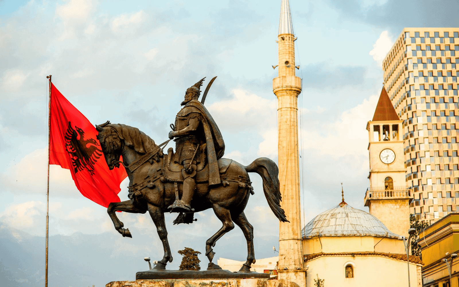 Statue of a man on a horse, the Albanian flag, and the clock tower of Skanderbeg Square in Tirana, Albania