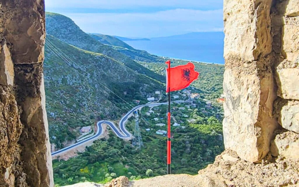 Vibrant red Albanian flag flying over scenic mountainous coastal landscape, emphasizing travel and exploration.