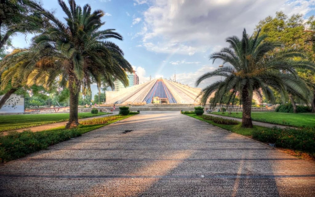 The Tirana Pyramid, one of the most famous attractions in Tirana, surrounded by palm trees and a blue sky with clouds