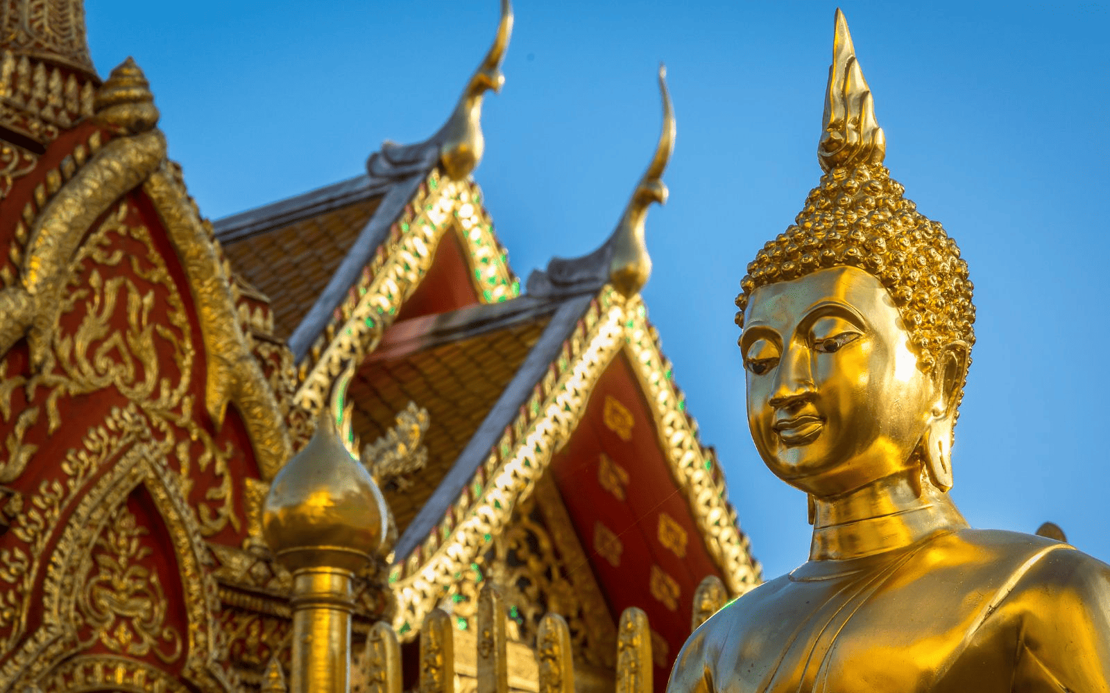 Gold and red Thai buddhis temple with golden buddha in front against a blue sky