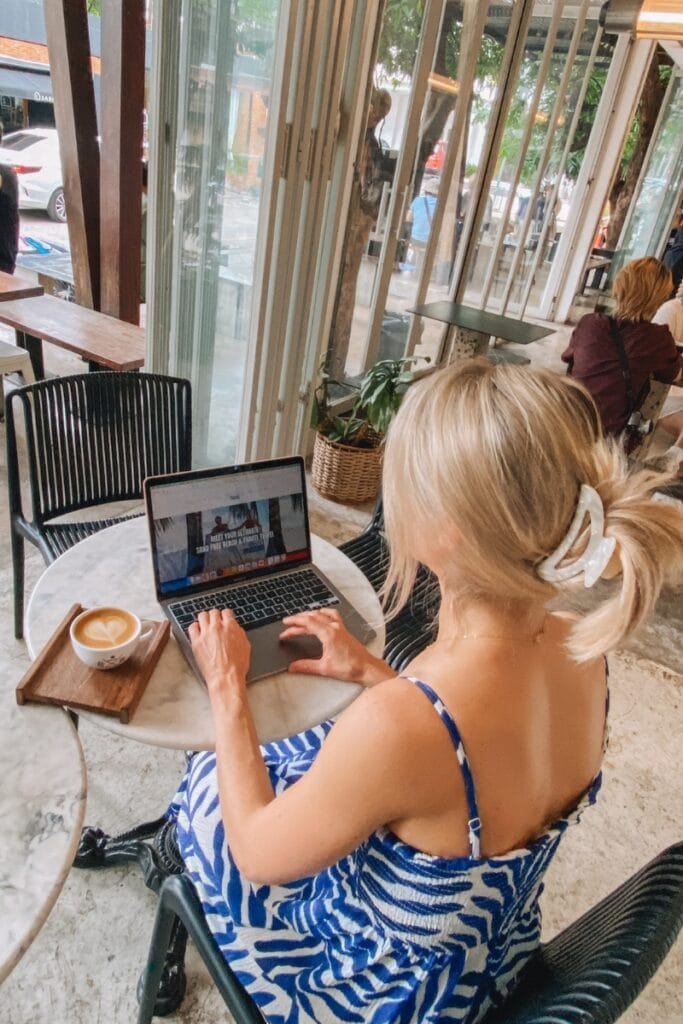 Relaxed woman working on a laptop at a cozy cafe, enjoying coffee and a scenic indoor-outdoor atmosphere.