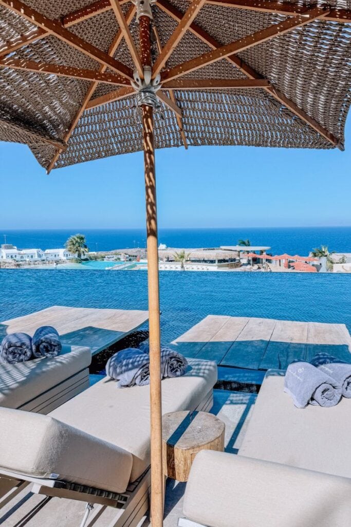 Poolside beach chairs under a woven umbrella with stunning sea and sky views in Santorini.