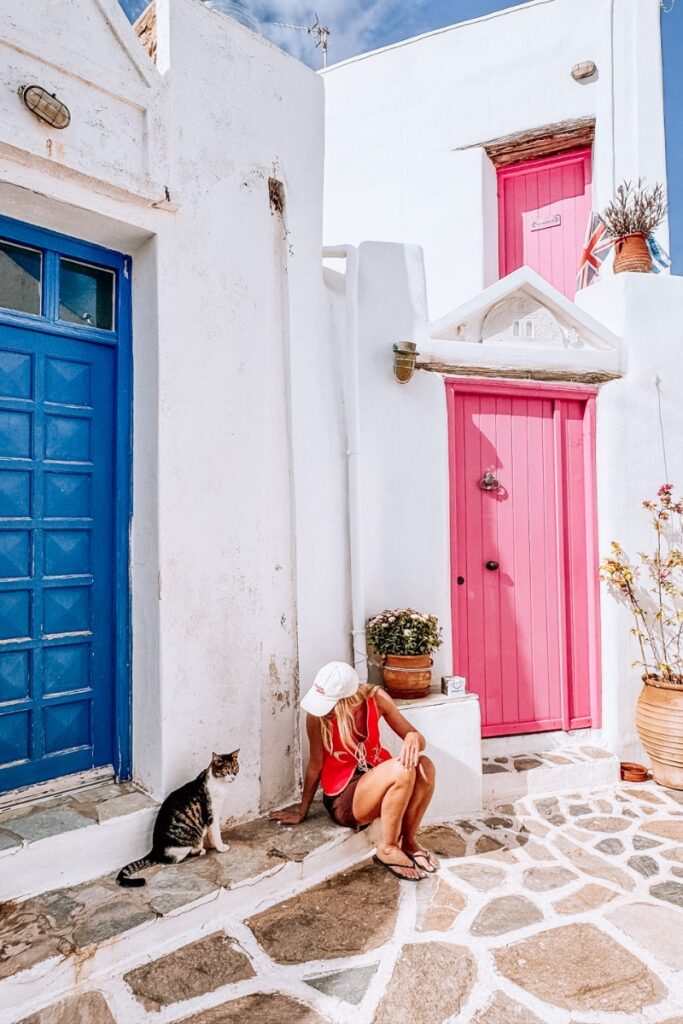 Woman sitting by vibrant pink door with cat in a Greek alley, white walls, and cobblestone street.