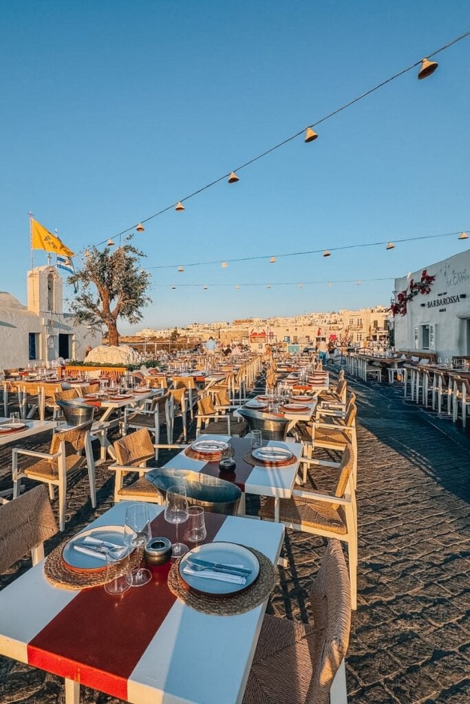 Outdoor Greek restaurant with beach view, wooden tables, and string lights.