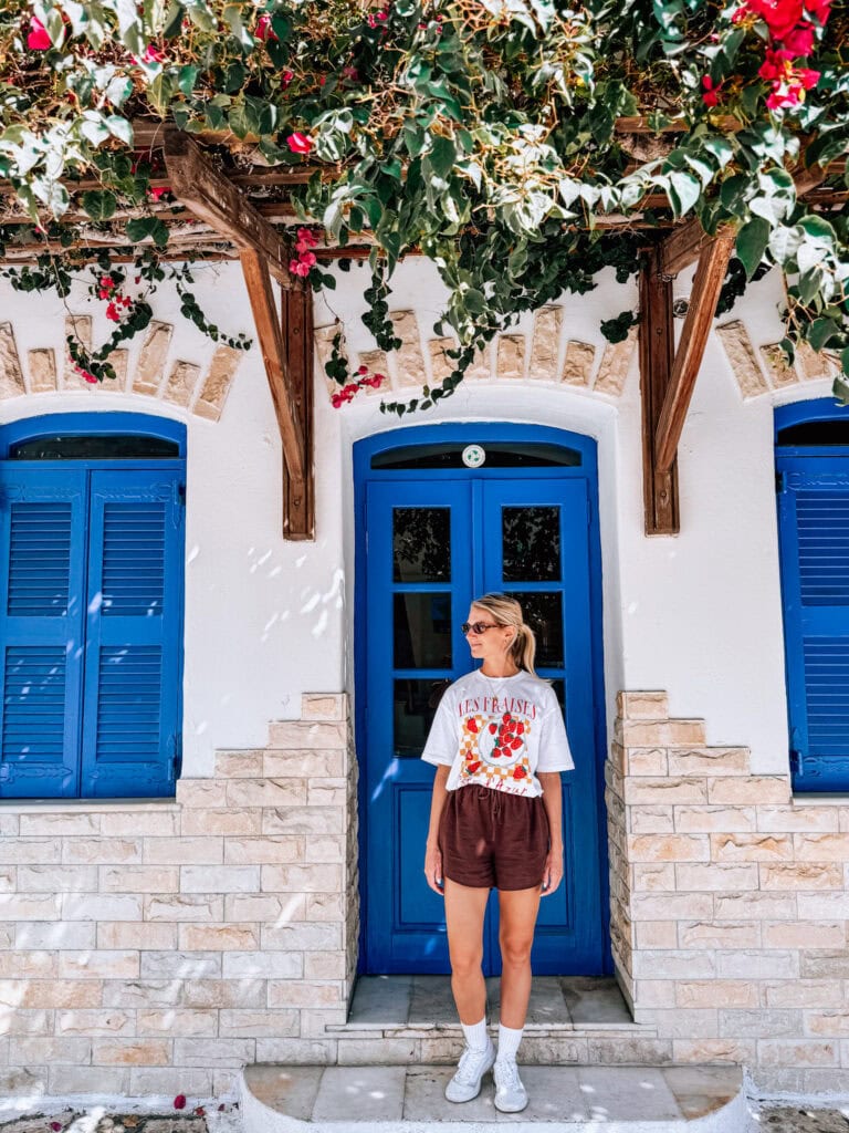 Girl standing in front of a traditional white and blue Greek house with lush greenery and flowers.