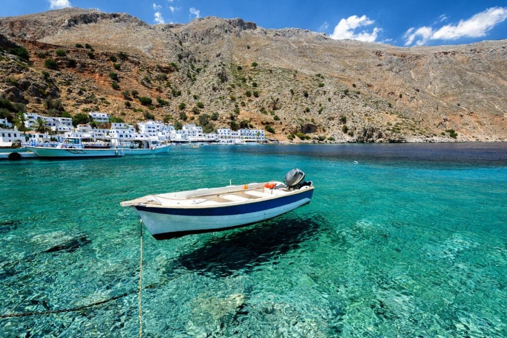 A boat floating on clear turquoise water near a coastal village with mountains in the background.